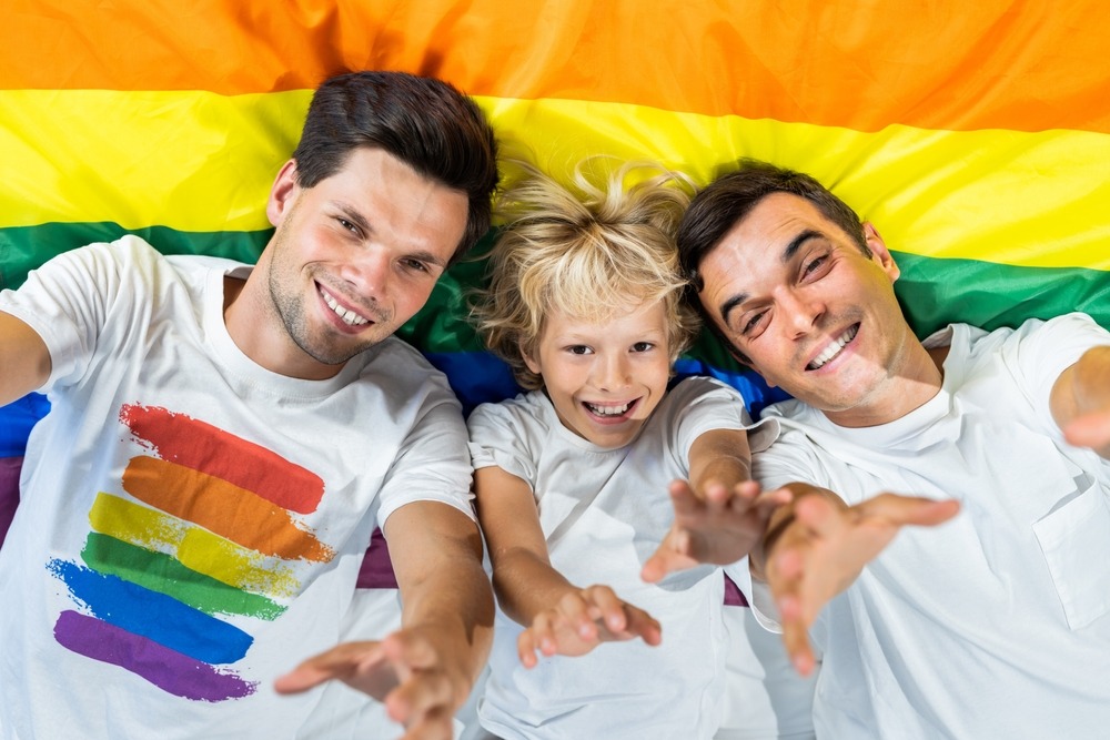 A family of two dads and a child laying on a rainbow flag.